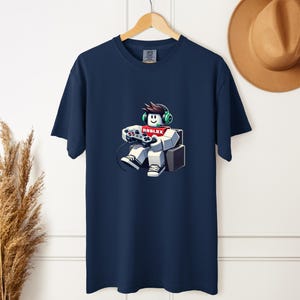 May include: Navy blue t-shirt featuring a cartoon character playing a video game. The character is wearing headphones and holding a game controller. The word "ROBLOX" is displayed on the character's shirt. The shirt is hanging on a wooden hanger.