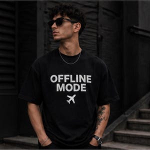 以下が含まれることがあります： 白文字で「OFFLINE MODE」と小さな飛行機のグラフィックがプリントされた黒いTシャツ。半袖でゆったりとしたフィット感です。
