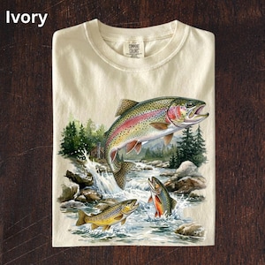 Puede incluir: Camiseta de color marfil con una vibrante ilustración de tres peces saltando de un río. El diseño incluye una trucha arcoíris, una trucha marrón y un pez de aleta roja, sobre un fondo de árboles y una pequeña cabaña. La camiseta tiene una etiqueta Comfort Colors.