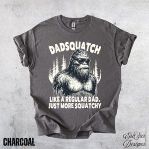 Può includere: T-shirt grigio antracite con la scritta "Dadsquatch" sopra una grafica di un Bigfoot con occhiali da sole. Il testo "Like a regular dad, just more squatchy" è sotto. La maglietta ha le maniche arrotolate.