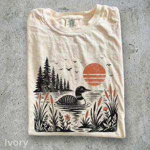 May include: An ivory t-shirt featuring a black and orange graphic print of a loon swimming on a lake. The design includes a forest of pine trees, cattails, a setting sun with horizontal lines, and birds flying in the sky. The inner label reads Comfort Colors.