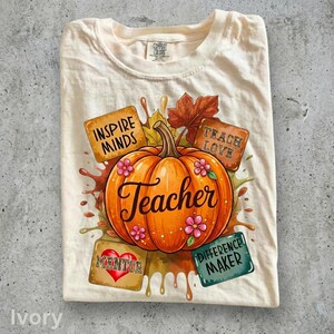 Op de afbeelding: Een ivoorkleurig t-shirt met een centrale oranje pompoenafbeelding met het woord Teacher in cursief schrift. Rondom de pompoen bevinden zich herfstbladeren en vier rechthoekige blokken met de tekst Inspire Minds, Teach Love, Mentor en Difference Maker.