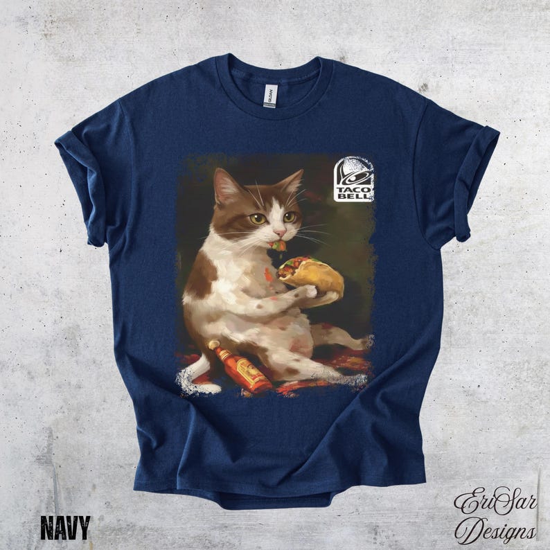 May include: Navy blue t-shirt featuring a cat eating a taco, with a Taco Bell logo. The cat is brown and white, with a taco in its paws. Two bottles of hot sauce are visible. The word "NAVY" is printed at the bottom.
