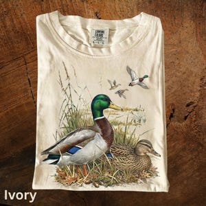 Könnte beinhalten: Elfenbeinfarbenes T-Shirt mit einer detaillierten Illustration von zwei Enten in einer natürlichen Umgebung, mit mehreren Enten im Flug. Das Shirt hat einen Rundhalsausschnitt und das Etikett der Marke Comfort Colors. Das Wort "Ivory" ist unten aufgedruckt.