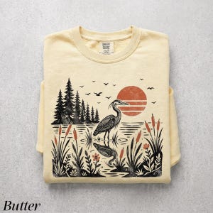 May include: A pale yellow crewneck sweatshirt featuring a graphic print of a heron standing in a marsh with cattails, pine trees, and a large orange sun. The design includes birds in flight and the text Comfort Colors on the inner neck label.