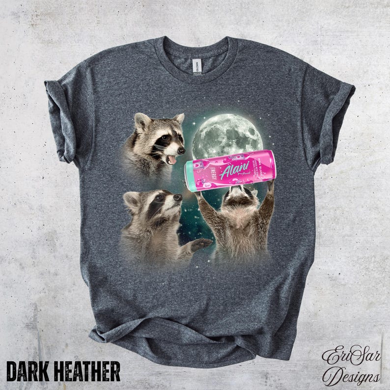 May include: Dark heather gray t-shirt featuring a whimsical design of three raccoons under a full moon. One raccoon holds a pink Alani Nu energy drink can. The shirt has the text "DARK HEATHER" at the bottom.