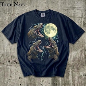 Könnte beinhalten: Ein dunkelblaues T-Shirt mit einem Grafikdruck von drei Komodowaranen mit geöffneten Mäulern, umgeben von hellblauen Blitzen und einem großen, leuchtend gelben Vollmond. Der Text TRUE NAVY ist oben links aufgedruckt.