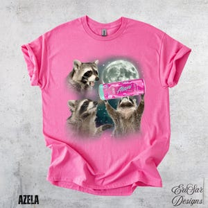 May include: A pink t-shirt featuring a graphic of three raccoons with a full moon in the background. One raccoon holds a pink Alani Nu energy drink. The shirt has the word "AZELA" printed at the bottom left.