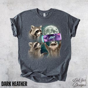May include: Dark heather gray t-shirt featuring three raccoons with a full moon background. One raccoon holds a can of Alani Nu energy drink. The shirt has the text "DARK HEATHER" at the bottom.
