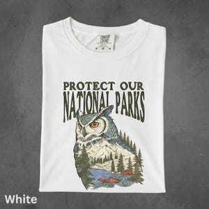 Peut inclure: T-shirt blanc avec l'inscription "PROTECT OUR NATIONAL PARKS" et une illustration d'un hibou avec un paysage de montagne et de for&ecirc;t. Le t-shirt est pli&eacute; proprement.