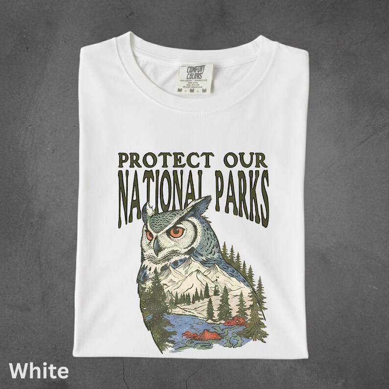 Peut inclure: T-shirt blanc avec l'inscription "PROTECT OUR NATIONAL PARKS" et une illustration d'un hibou avec un paysage de montagne et de for&ecirc;t. Le t-shirt est pli&eacute; proprement.