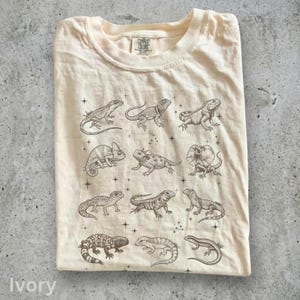 Może przedstawiać: Kremowy t-shirt z ciemnobrązowym nadrukiem graficznym przedstawiającym dwanaście różnych jaszczurek ułożonych w siatkę. Wzór zawiera różne gatunki, takie jak kameleon, agama kołnierzasta i gekon, przeplatane małymi ikonami gwiazdek.