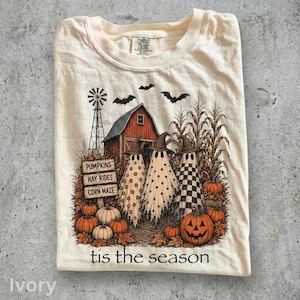 May include: An ivory t-shirt featuring a graphic of three ghosts wearing witch hats in front of a red barn, a windmill, and corn stalks. The print includes pumpkins, bats, and signs reading PUMPKINS, HAY-RIDES, CORN MAZE, with the text tis the season.