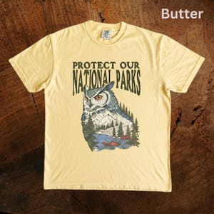Peut inclure: T-shirt jaune beurre avec l'inscription "PROTECT OUR NATIONAL PARKS" et une illustration de hibou. Le hibou est repr&eacute;sent&eacute; avec une sc&egrave;ne de montagne et de for&ecirc;t. Le hibou a des plumes bleues, blanches et grises.