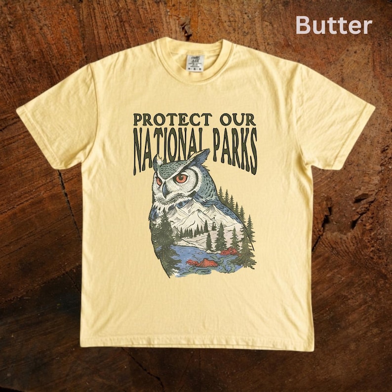 Peut inclure: T-shirt jaune beurre avec l'inscription "PROTECT OUR NATIONAL PARKS" et une illustration de hibou. Le hibou est repr&eacute;sent&eacute; avec une sc&egrave;ne de montagne et de for&ecirc;t. Le hibou a des plumes bleues, blanches et grises.