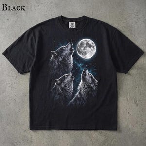 May include: A black short-sleeve t-shirt featuring a graphic print of three wolves howling at a full moon. The design includes a starry night sky background with blue nebula-like accents. The shirt has a crew neckline and a standard fit.