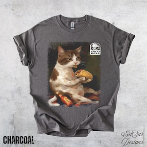 May include: Charcoal-colored t-shirt featuring a whimsical illustration of a cat enjoying a taco, with a Taco Bell logo. The cat is brown and white, holding a taco and a bottle of hot sauce. The text "CHARCOAL" is at the bottom.