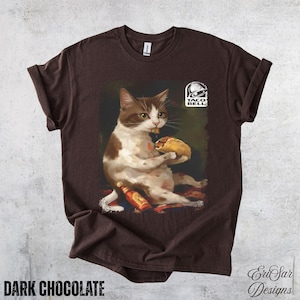 May include: Dark chocolate brown t-shirt featuring a cat holding a taco and a bottle of hot sauce. The cat is brown and white, and the Taco Bell logo is visible. The text "DARK CHOCOLATE" is at the bottom of the shirt.