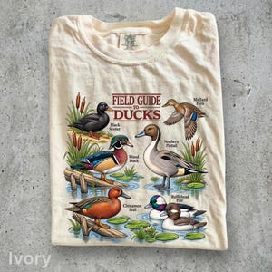 Könnte beinhalten: Ein cremefarbenes T-Shirt mit einer Grafik mit der Aufschrift Field Guide to Ducks. Die Illustration zeigt eine Trauerente, eine Brautente, eine Spießente, eine Stockente und ein Büffelkopf-Paar in einem Teich mit Schilf und Baumstämmen.