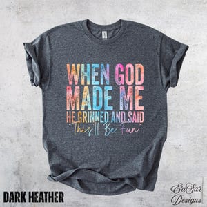 Op de afbeelding: Donkergrijs heather t-shirt met de tekst "WHEN GOD MADE ME HE GRINNED AND SAID 'This'll Be Fun'" in een kleurrijk lettertype in aquarel-stijl. De tekst "DARK HEATHER" staat er ook op.