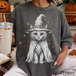 May include: A charcoal gray short-sleeve t-shirt featuring a white graphic illustration of a cat wearing a pointed witch hat and a cape. The design is accented with small yellow stars scattered around the cat, set against a solid dark background.