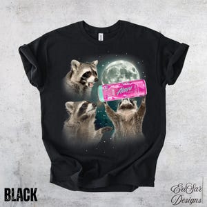 May include: Black t-shirt featuring a graphic of three raccoons with a full moon in the background. One raccoon holds a pink can of "Alani" energy drink. The word "BLACK" is printed at the bottom.