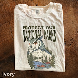 May include: Ivory-colored t-shirt with the text "PROTECT OUR NATIONAL PARKS" above an owl illustration. The owl design incorporates a mountain and forest landscape in shades of blue, green, and brown. The shirt is folded on a wooden surface.