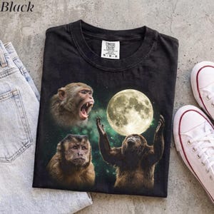 Può includere: Una t-shirt nera con una stampa grafica di tre scimmie su uno sfondo spaziale verde scuro con una grande luna piena luminosa. Una scimmia ha la bocca aperta, un'altra guarda avanti e la terza tende le braccia verso la luna.