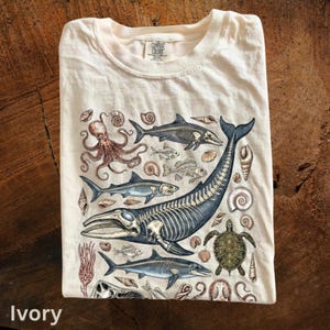 以下が含まれることがあります： アイボリー色のTシャツ。クジラの骨格、サメ、タコ、貝殻など、さまざまな海洋生物の詳細なイラストが描かれています。デザインは青、茶色、クリーム色を使用し、ヴィンテージ感を演出しています。シャツはきれいに折りたたまれています。