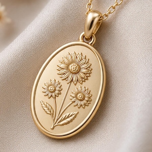 May include: A gold-toned oval pendant necklace featuring an embossed design of three sunflowers with stems and leaves. The pendant hangs from a delicate matching gold-toned chain, showcasing a smooth, polished finish on the outer rim and bail.