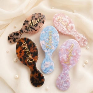 Op de afbeelding: Vijf gepersonaliseerde haarborstels in verschillende kleuren en patronen. De ovale borstels met handvatten hebben gouden namen: Alice, Charlotte, Eleanor, Seraphina en Beatrix. De achtergrond is een zachte, witte stof.