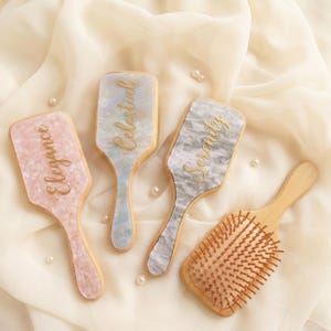 May include: Four hairbrushes with wooden handles and different brush heads. Three have decorative tops with the words "Elegance", "Celestial", and "Serenity" in gold script. The fourth brush has wooden bristles.