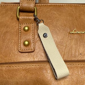 May include: Close-up of a tan leather handbag with a cream leather keychain attached. The handbag has gold-coloured hardware, including rivets and a clasp. The keychain is secured with a silver-coloured metal clasp.