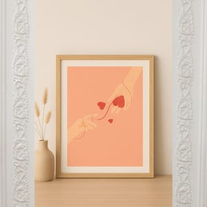 Könnte beinhalten: Gerahmter Kunstdruck mit einer Illustration von zwei Händen, die sich einander entgegenstrecken, mit roten Herzen, die zwischen ihnen schweben. Das Kunstwerk hat einen minimalistischen Stil mit einem pfirsichfarbenen Hintergrund und einem Holzrahmen. Links eine Vase mit Trockenblumen.