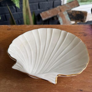 May include: A white ceramic dish shaped like a scallop shell, featuring a smooth, glossy interior with radiating ridges and a thin, metallic gold-colored trim along the scalloped outer edge, designed as a decorative bowl or trinket tray.