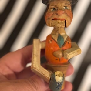 May include: A hand-carved wooden nutcracker figure wearing a black hat, an orange jacket with a blue tie, and light-colored trousers. The figure features a painted face with defined facial features and a movable jaw mechanism for cracking nuts.