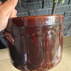 May include: A cylindrical brown ceramic crock featuring a glossy glaze and a decorative scalloped relief pattern around the upper rim. The vessel includes two small side handles and exhibits a deep, dark brown finish with subtle variations in tone.