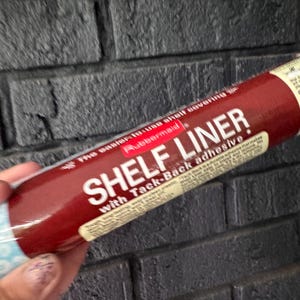 May include: A cylindrical roll of Rubbermaid shelf liner with a dark red label featuring white text that reads SHELF LINER with Tack-Back adhesive. The packaging includes additional descriptive text and a light blue patterned section at the bottom.