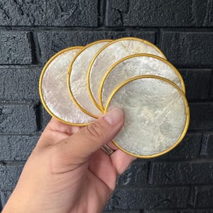 May include: A set of five circular coasters featuring a shimmering, iridescent white mother-of-pearl inlay surface. Each coaster is framed by a thin, solid gold-colored metal rim, creating a clean and uniform appearance for this collection of drink accessories.