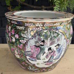 May include: A round ceramic planter featuring a colourful hand-painted scene of four figures in traditional robes within a central panel. The exterior is decorated with gold accents, pink flowers, and green foliage against a dark background.