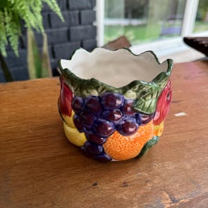 May include: A ceramic bowl with a scalloped rim featuring a three-dimensional relief design of colorful fruits including purple grapes, a bright orange, and a red apple. The interior of the bowl is smooth and white, while the exterior is textured.