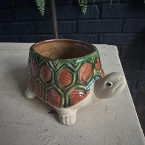 May include: A ceramic turtle-shaped planter featuring a glossy finish with a green and orange shell pattern. The turtle has a light beige head, four feet, and a hollow interior for planting. The design includes textured hexagonal shell segments.