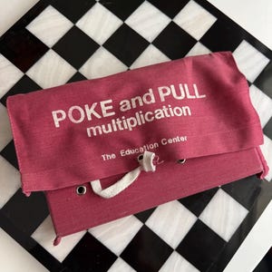 May include: A dusty rose fabric educational game pouch with a fold-over flap. The text POKE and PULL multiplication and The Education Center are printed in white. A white fabric tie is threaded through metal grommets on the front.