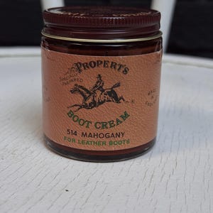 May include: A glass jar of Propert's boot cream with a dark brown metal lid. The tan label features a black illustration of a person riding a horse, with text reading Propert's Specially Prepared Boot Cream 514 Mahogany For Leather Boots.