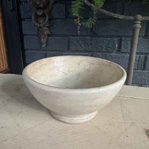 May include: A round, off-white ceramic bowl with a pedestal base. The bowl features a visible crackle glaze finish across its entire surface, showing subtle variations in tone and texture throughout the exterior and interior of the vessel.