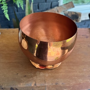 May include: A polished copper bowl with a rounded, spherical body and a small circular base. The exterior surface is reflective and smooth, showcasing a warm metallic orange-brown hue, while the interior reveals a slightly darker copper tone.