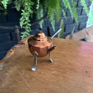 May include: A miniature copper teapot with a rounded body, a small lid featuring a decorative finial, and a short spout. The teapot is supported by three slender, silver-toned metal legs that extend outward from the base of the copper vessel.