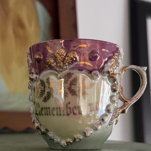 Puede incluir: Una pequeña y ornamentada taza de té con borde morado y detalles dorados. La taza presenta un diseño decorativo de cuentas y la palabra "Remember" en letras doradas. El asa y el ribete son plateados.