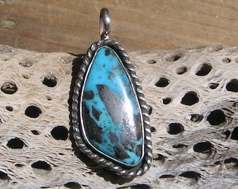 May include: A silver pendant featuring a blue and black turquoise stone. The pendant is set in a silver bezel and has a rope-like border.
