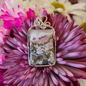 May include: A rectangular pendant with a silver wire wrap. The pendant features a stone with a white, pink, and green pattern. The pendant is set against a backdrop of pink and white flowers.